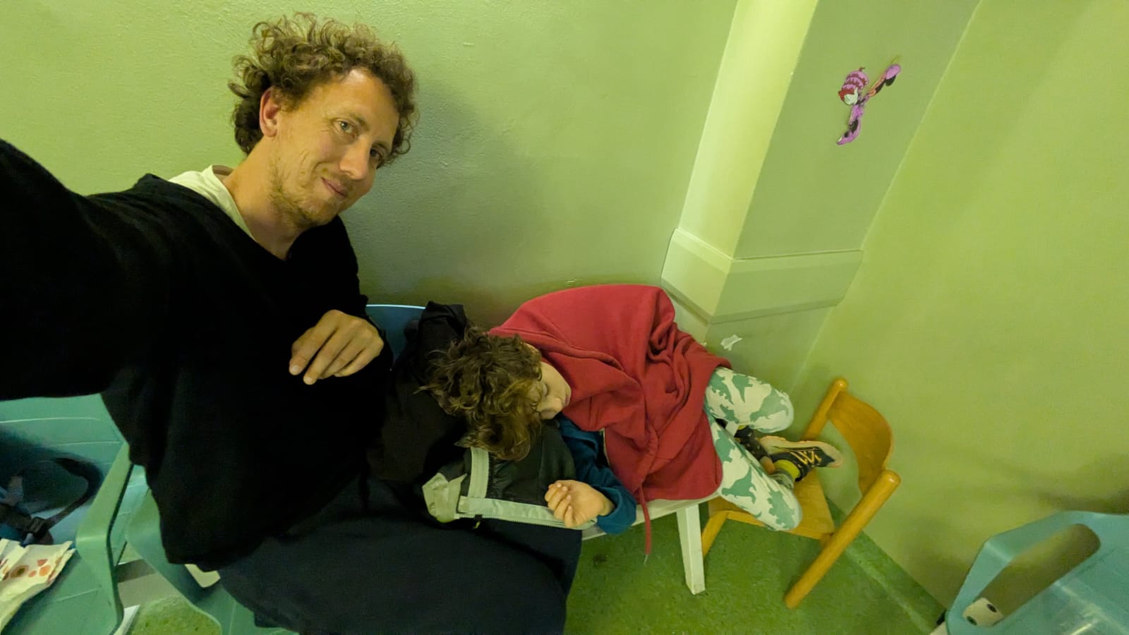 Marco e Diego nella sala d'attesa del pronto soccorso del Buzzi, nel cuore della notte: Marco seduto con il telefono sollevato per scattare il selfie, Diego raggomitolato accanto a lui sulla sedia, addormentato sotto una felpa rossa.